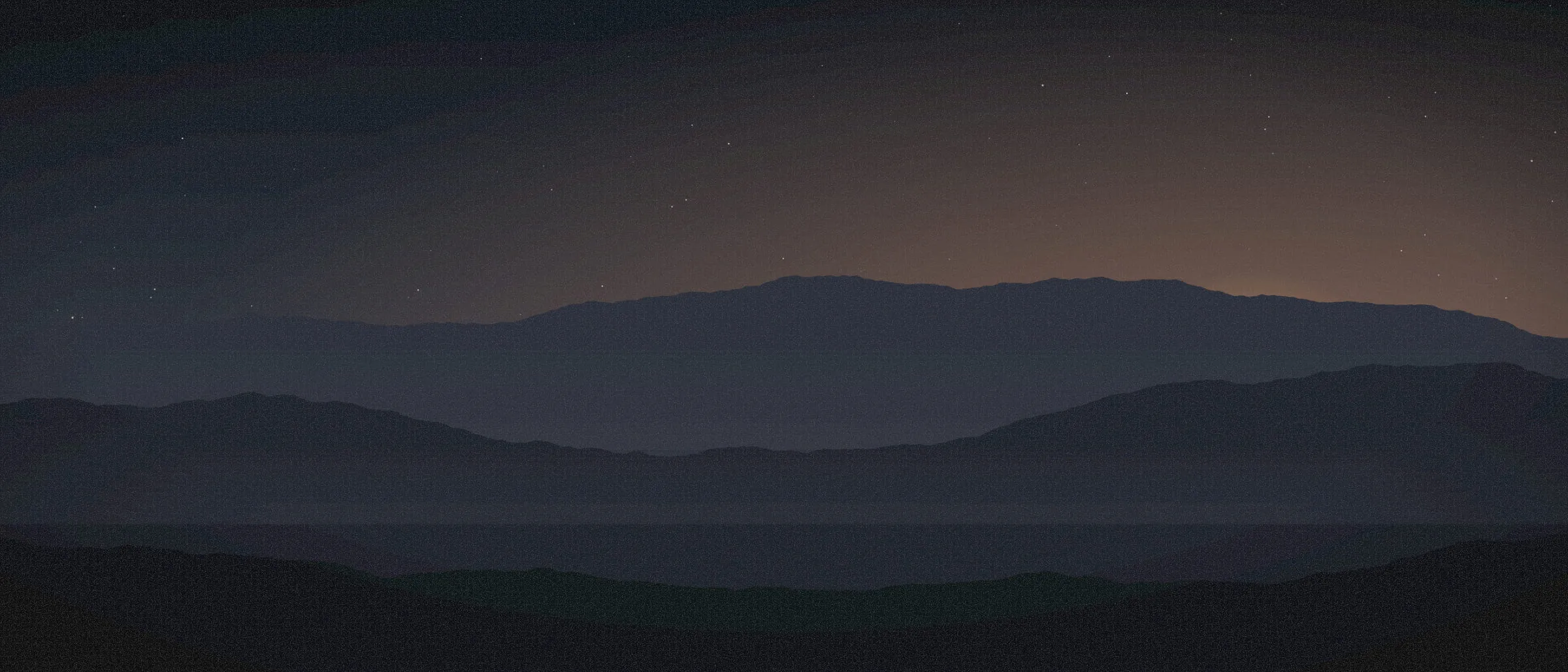 A wide panorama of overlapping blue ridges, an orange sun setting behind the farthest.